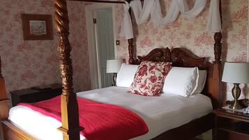 A four-poster bed with a red throw blanket, white linens, and floral pillows. There are two bedside tables with lamps. The room has floral wallpaper and a wooden ceiling.