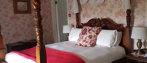 Una cama con dosel con un plaid rojo, sábanas blancas y almohadas estampadas. Hay dos mesitas de noche con lámparas. La habitación tiene papel tapiz floral y techo de madera.
