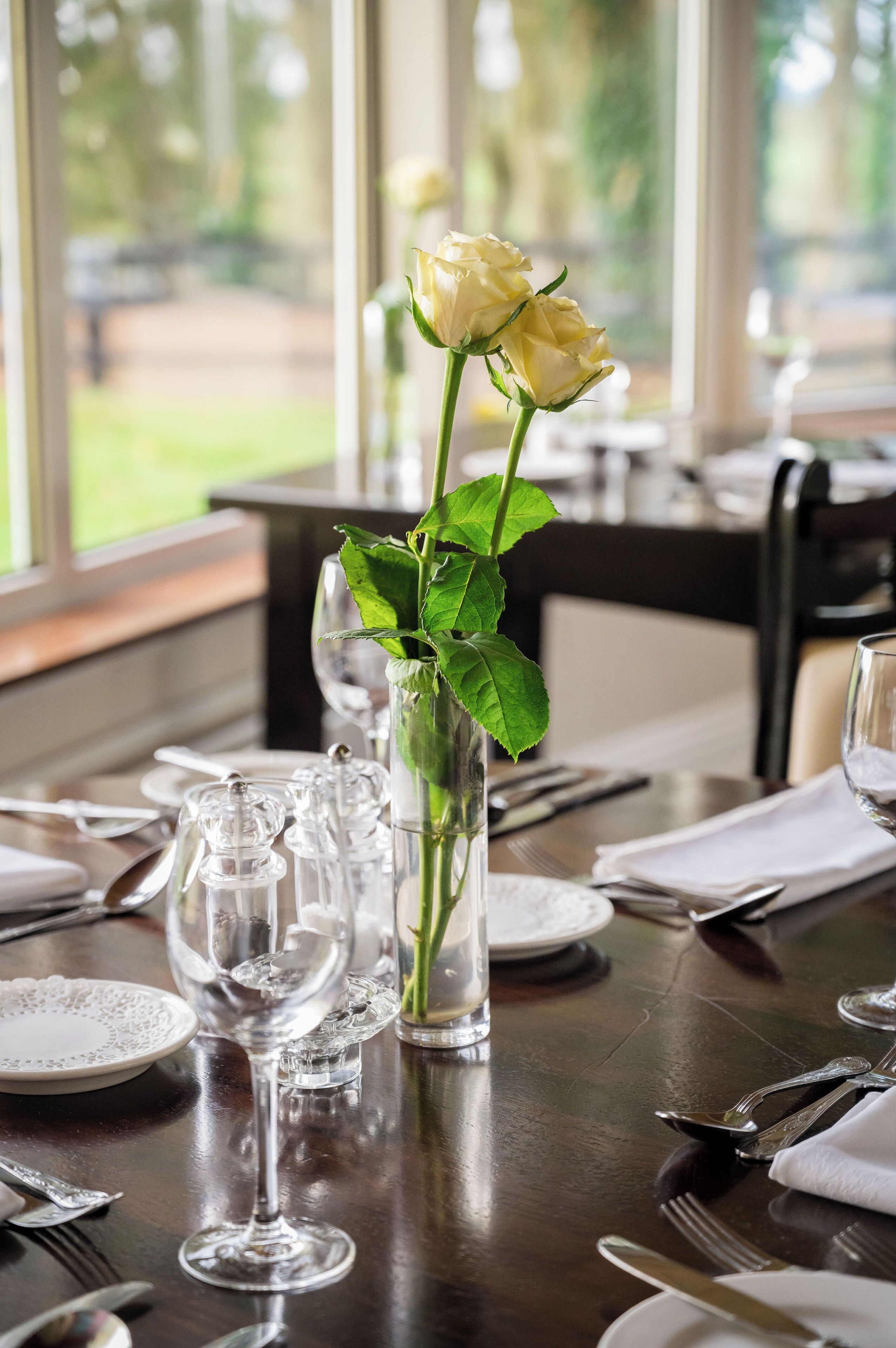 Une table à manger dressée avec de beaux verres transparents, une rose blanche dans un vase et une vue sur la verdure à l’extérieur.