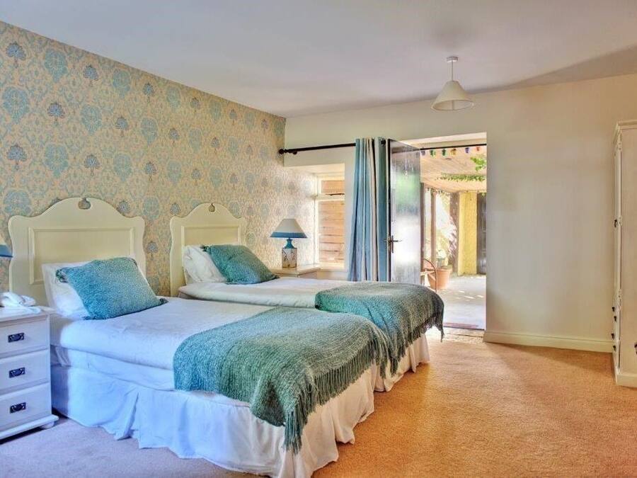 A bedroom with two beds, a nightstand, a lamp, and a view of the outdoors.