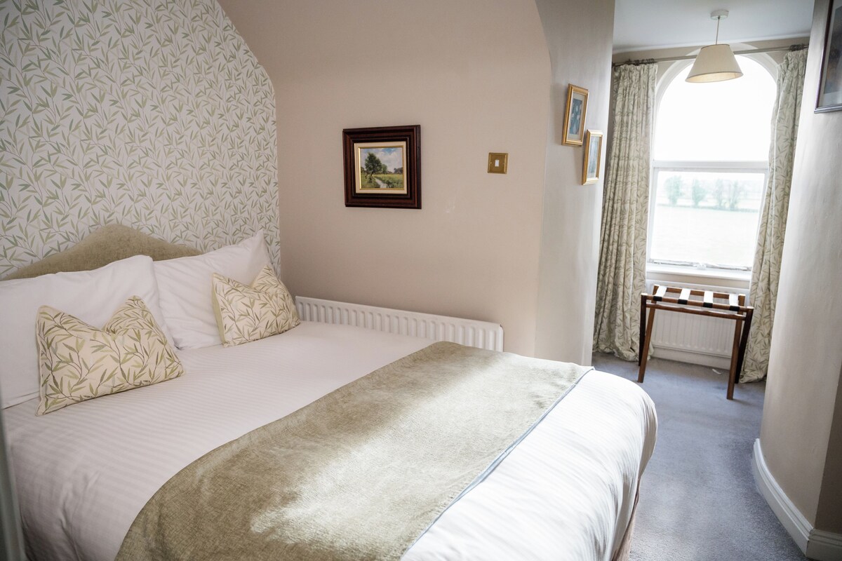 A neatly made bed with a white and beige duvet, a wooden bedside table, and a window with curtains.