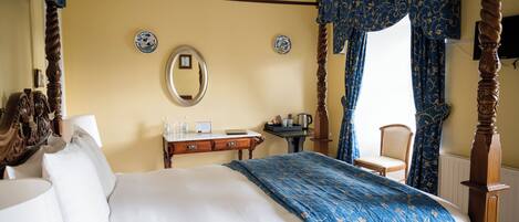 A four-poster bed with a blue floral bedspread, a bedside table with a mirror, a small table with a kettle, and a chair by the window.
