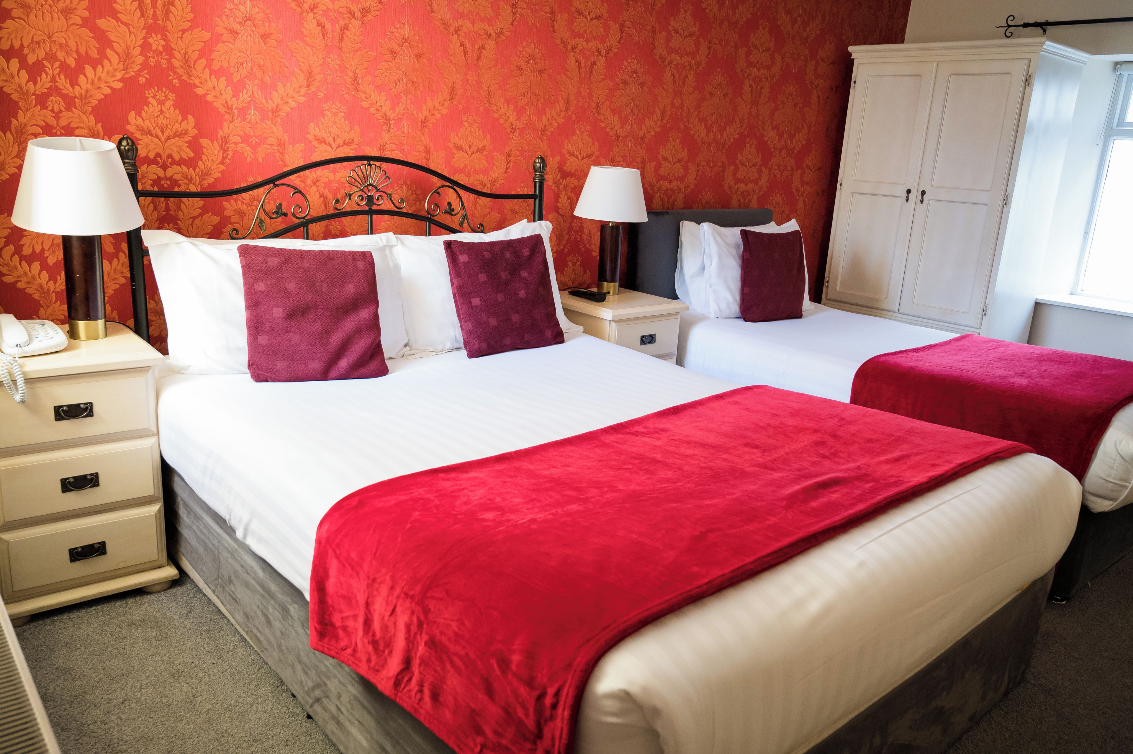 A hotel room with two beds, each with a red blanket, white pillows, and a nightstand with a lamp. There is a wardrobe and a window with curtains.