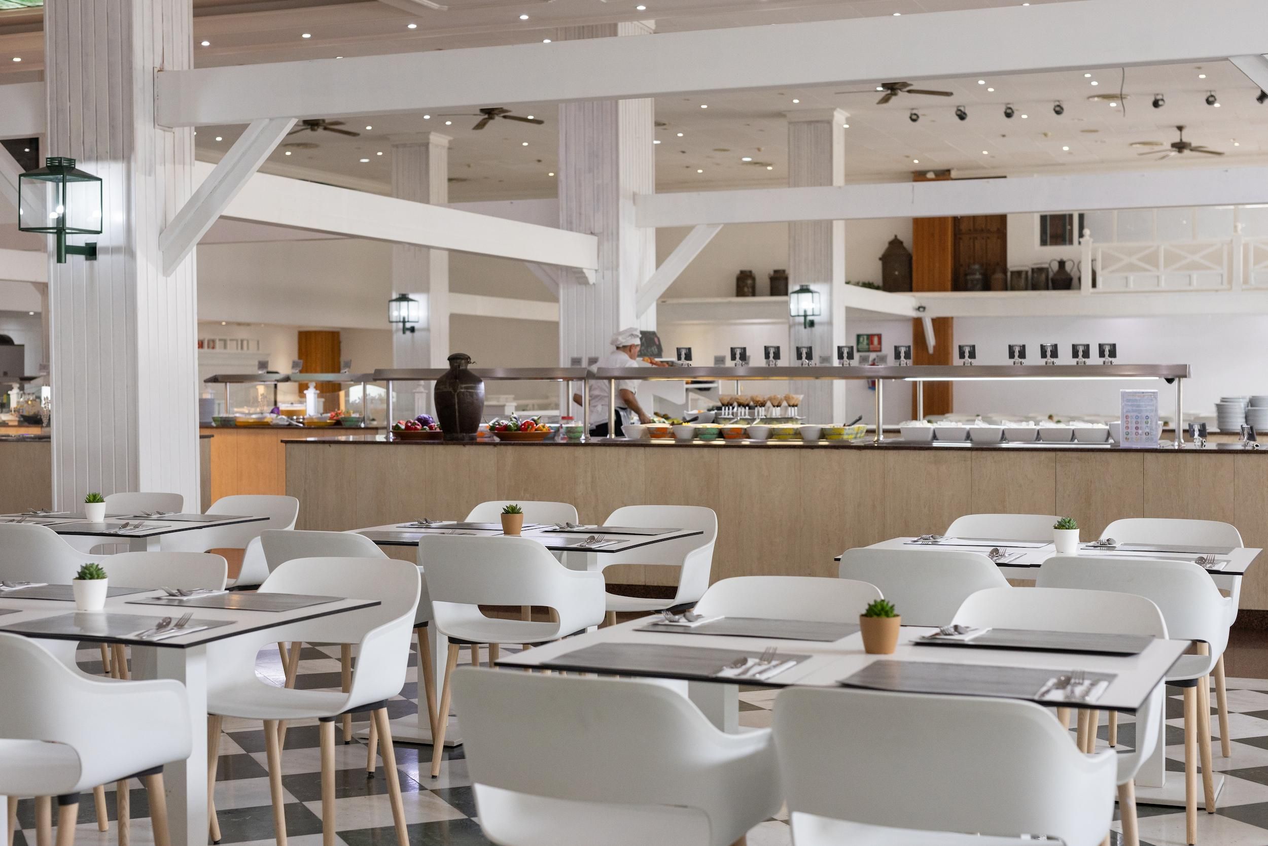 A modern restaurant with white chairs and tables, a central bar area with a chef, and a checkered floor.