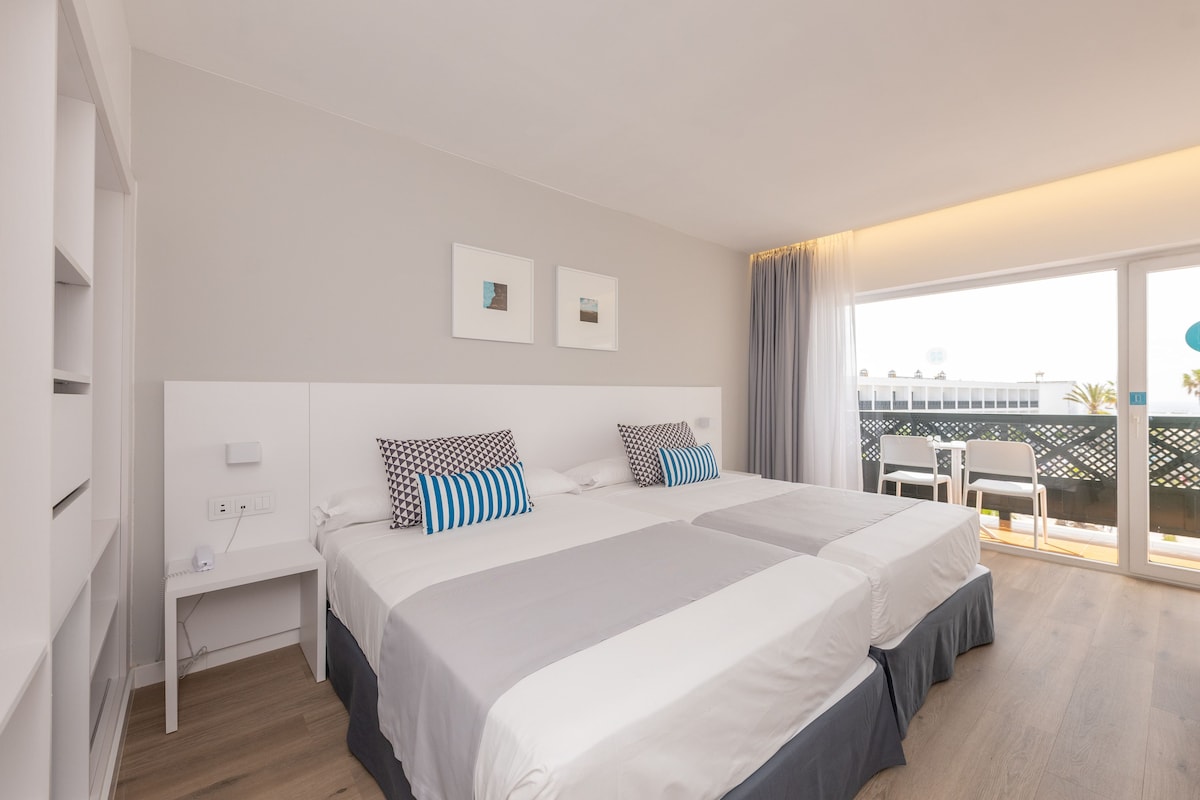 A hotel room with a large bed, two bedside tables, a window with curtains, and a view of the outside.