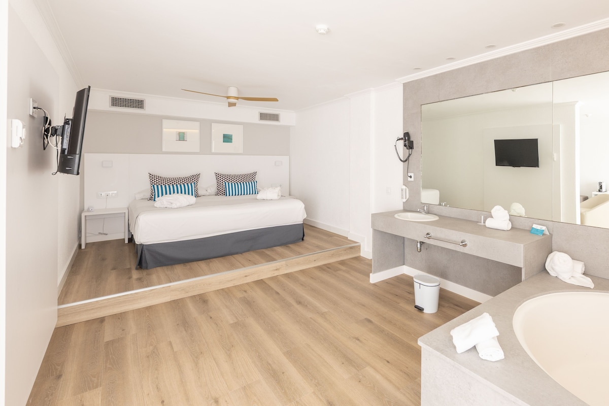 A modern hotel room with a bed, bedside tables, a large mirror, a vanity with a sink, and a shower area.