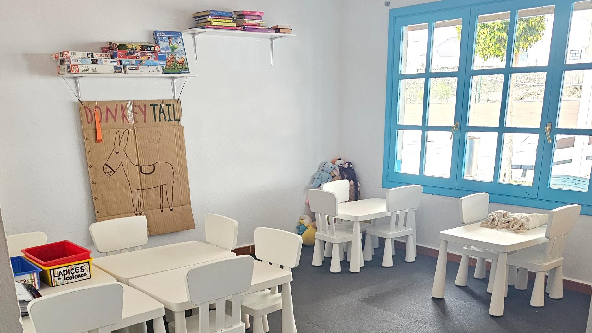 Un aula con sillas y mesas blancas, un ventanal con marcos azules y estanterías llenas de libros y juguetes.