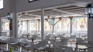 A modern restaurant with white chairs and tables, large windows with a view of palm trees, and a checkered floor pattern. - BLUESEA Costa Bastian (Teguise)