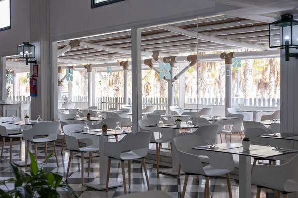 A modern restaurant with white chairs and tables, large windows with a view of palm trees, and a checkered floor pattern. - BLUESEA Costa Bastian (Teguise)