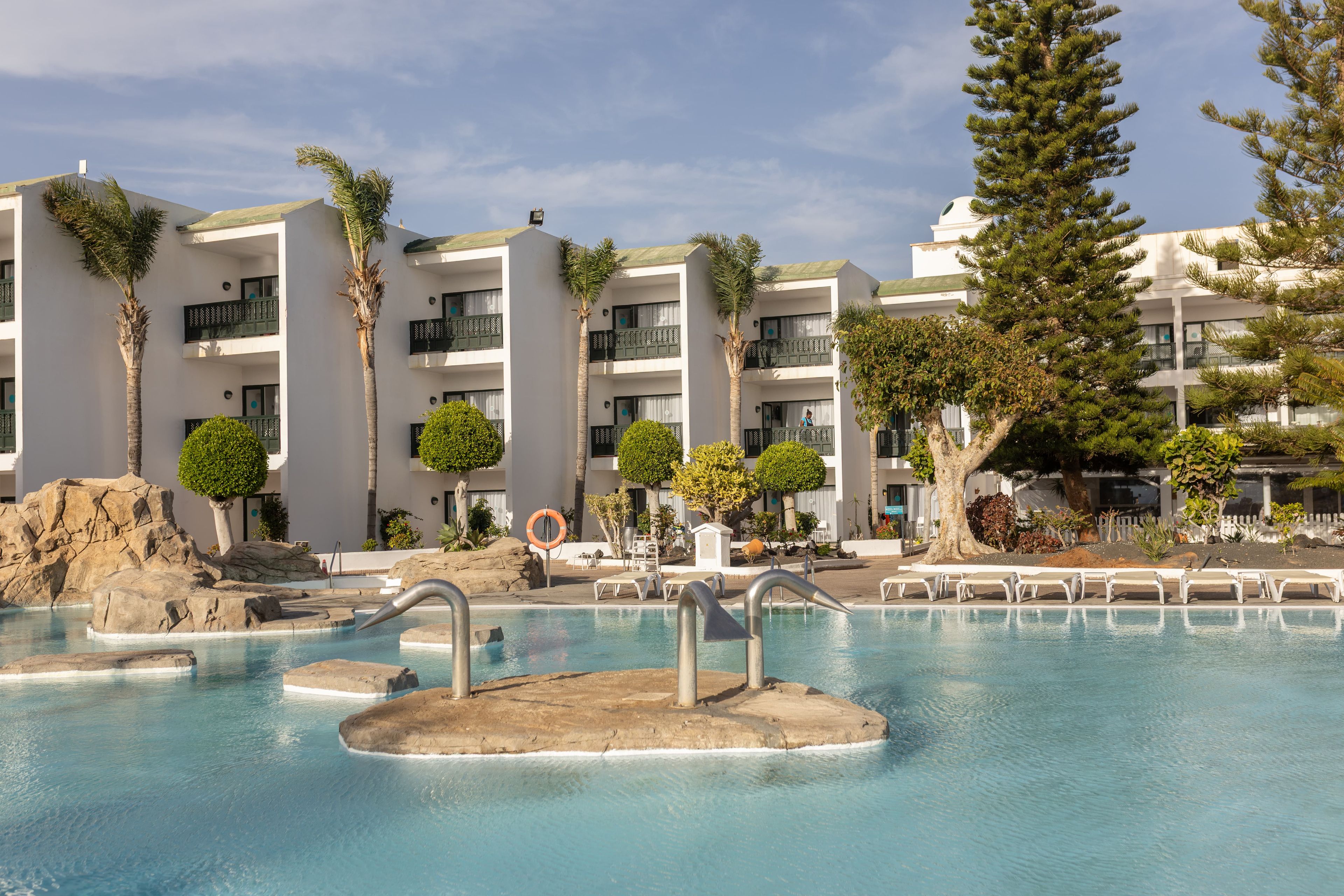 Un resort con área de piscina, palmeras y un edificio blanco con balcones.