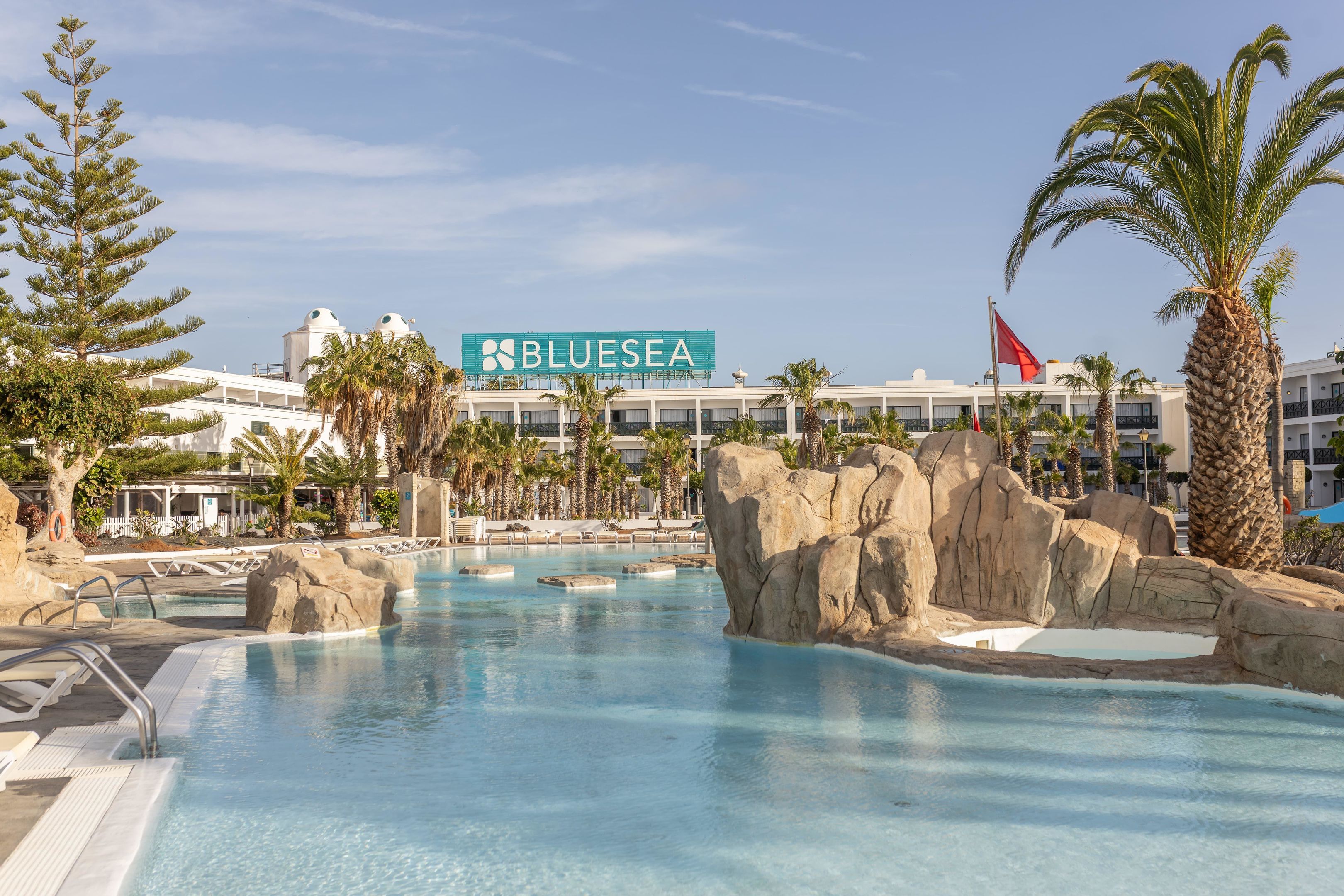 Zona de piscina en un resort con un letrero grande de BluSea, palmeras y una bandera.