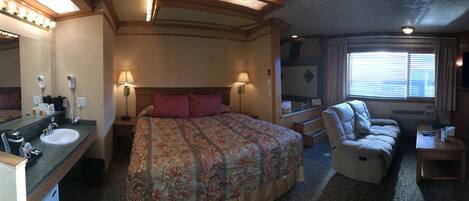 A hotel room with a large bed, a sitting area with a chair, a vanity with a mirror and sink, and a television mounted on the wall.