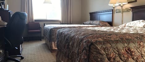 A hotel room with two beds, a desk, a chair, and a lamp.