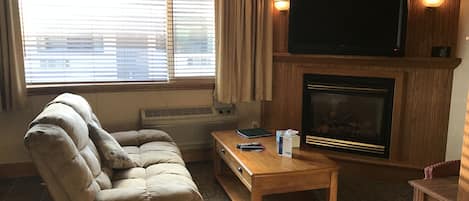 A living room with a beige sofa, a wooden coffee table, a flat-screen TV mounted above a fireplace, and a window with blinds.