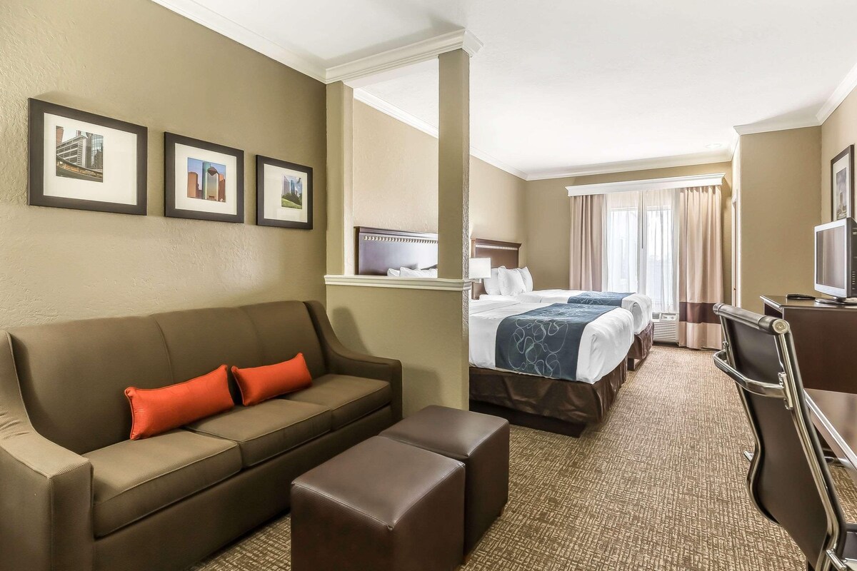 A hotel room with a sofa, ottoman, two beds, a desk, and framed pictures on the wall.