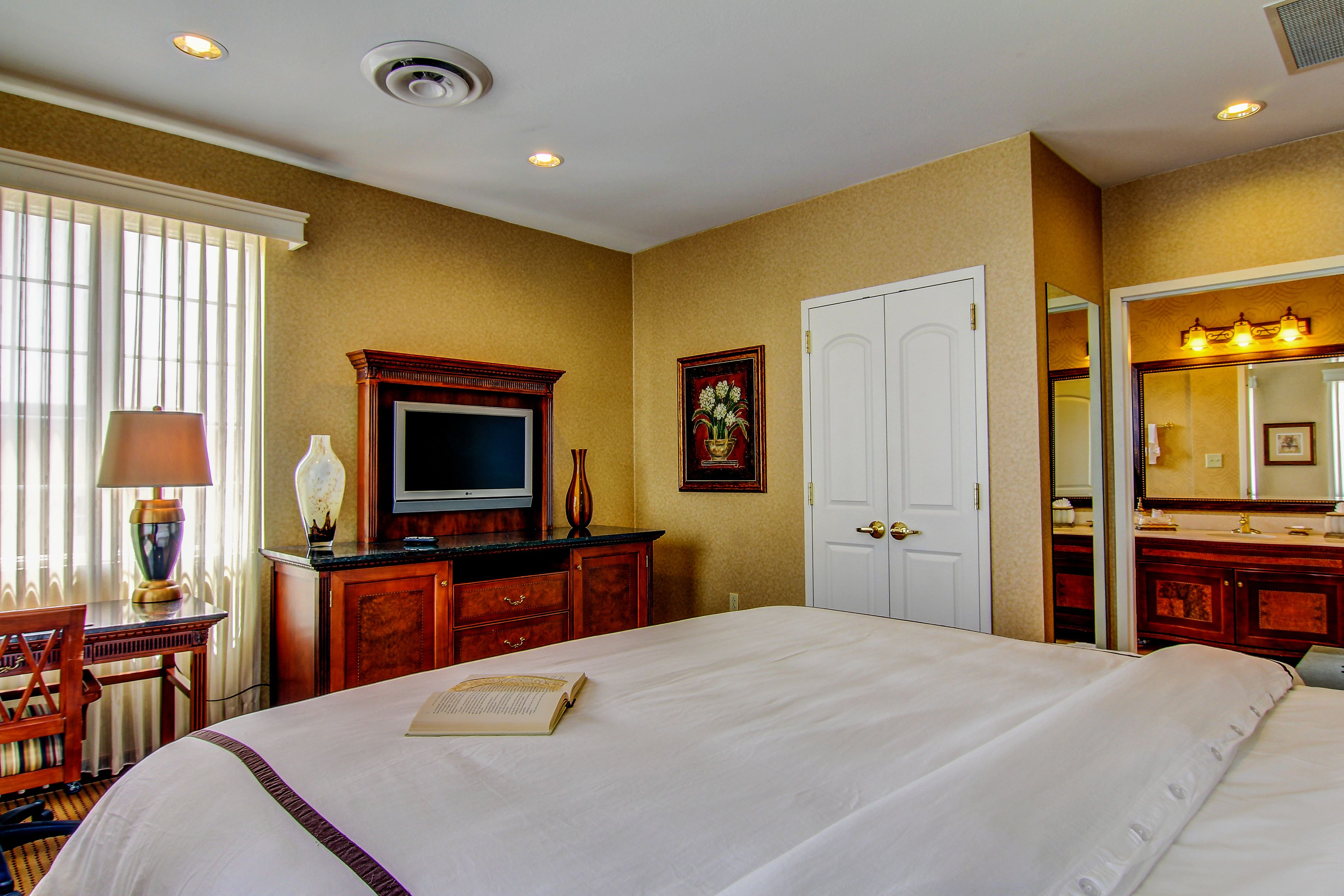 A hotel room with a large bed, a television, a desk with a lamp, a dresser, and a bathroom with a mirror and sink.