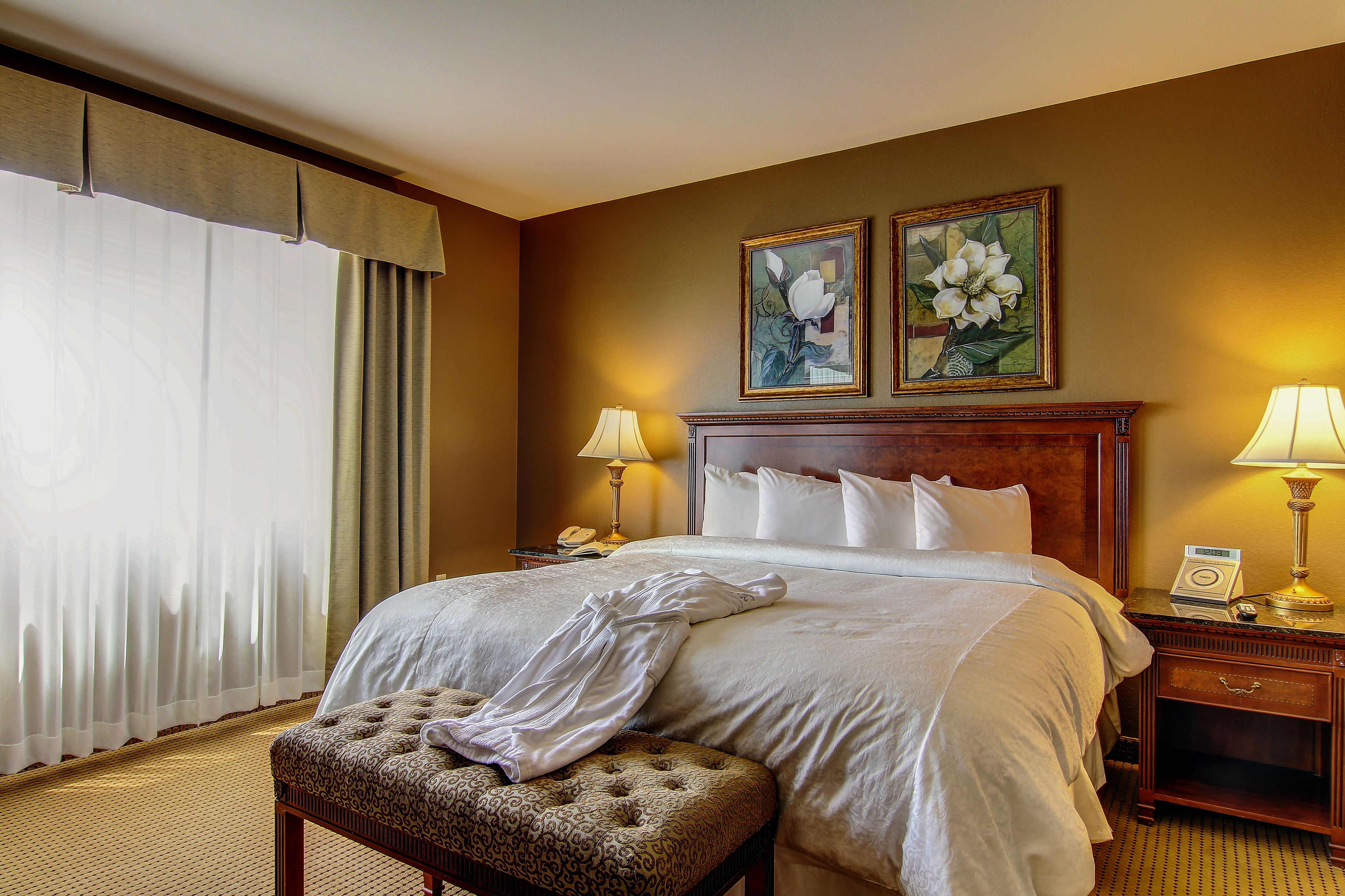 Una cama bien hecha con cubrecama y almohadas blancas, una mesita de noche de madera con una lámpara, una obra de arte floral enmarcada sobre la cama y una ventana con cortinas translúcidas.