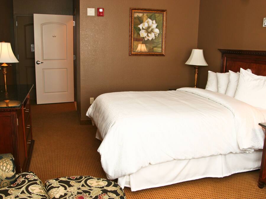 A hotel room with a large bed, two bedside lamps, a wooden headboard, a framed floral artwork on the wall, and a door leading to another room.