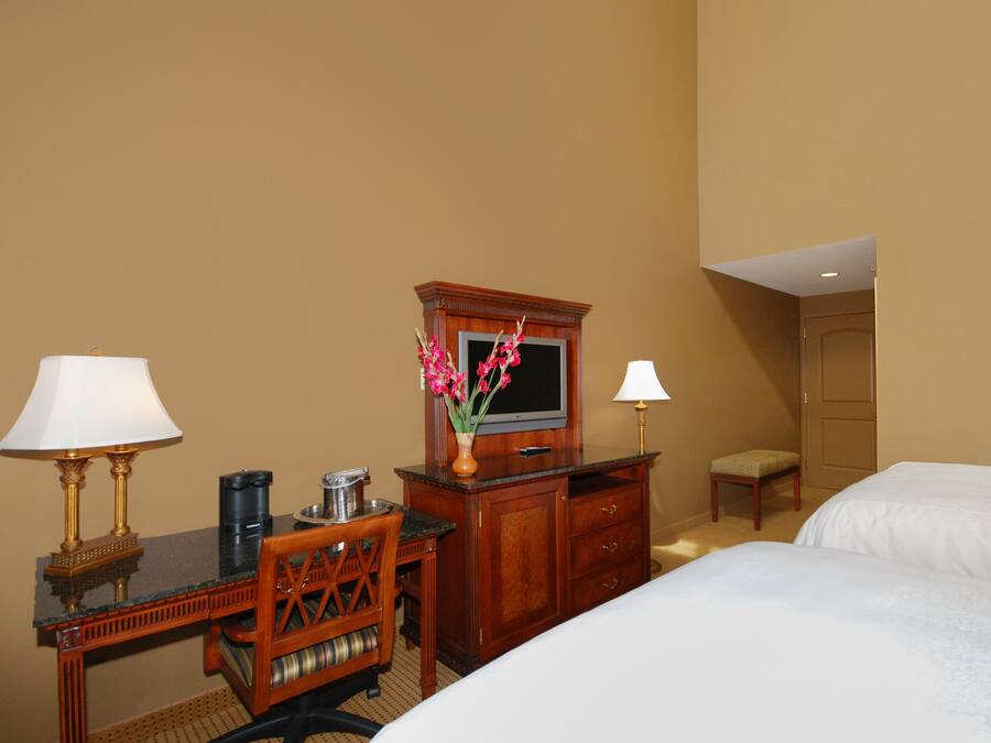 Ein Hotelzimmer mit Schreibtisch, zwei Lampen, Fernseher, Kommode und zwei Betten.