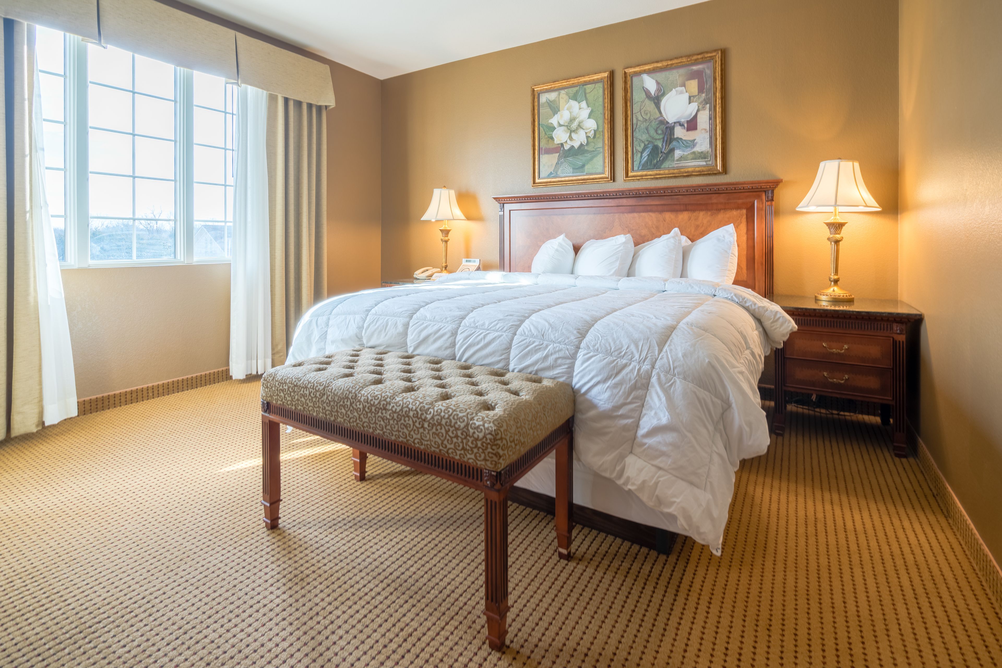 A hotel room with a large bed, two bedside lamps, a wooden nightstand, a bench, and two framed floral paintings on the wall.
