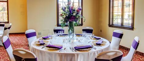 Une table ronde recouverte d’une nappe blanche, ornée de rubans violets et agrémentée d’un vase de fleurs.