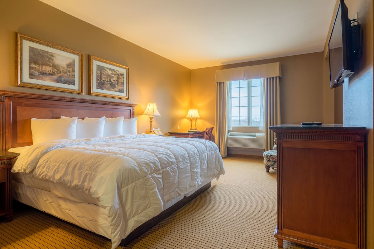 A hotel room with a large bed, two bedside lamps, a wooden nightstand, a desk, a chair, a dresser, a flat-screen TV, and two framed pictures on the wall.
