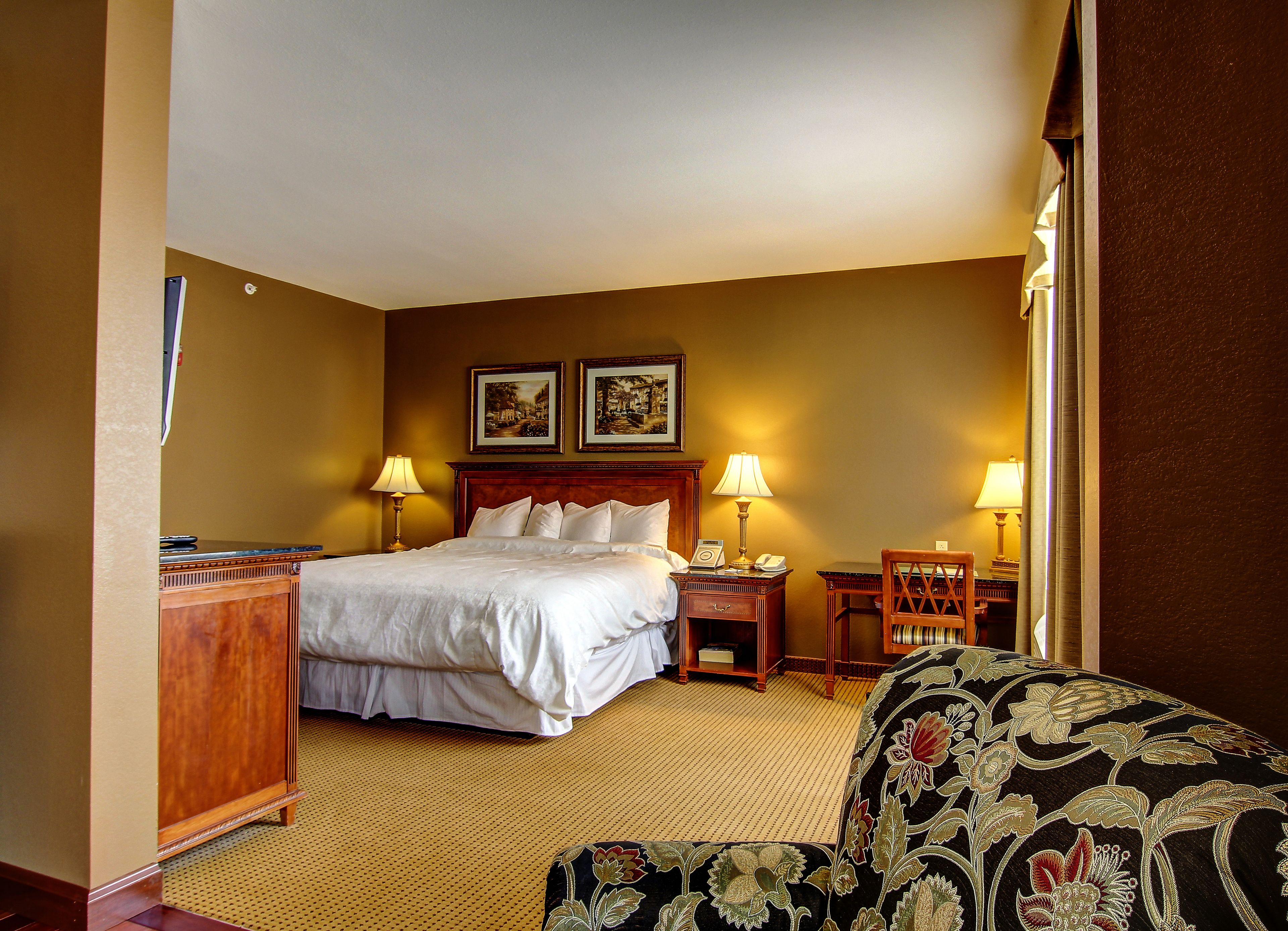 A hotel room with a large bed, two bedside lamps, a wooden nightstand, a desk, and a patterned armchair.