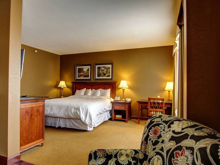 Ein Hotelzimmer mit einem großen Bett, zwei Nachttischlampen, einem Holznachttisch, einem Schreibtisch und einem gemusterten Sessel.