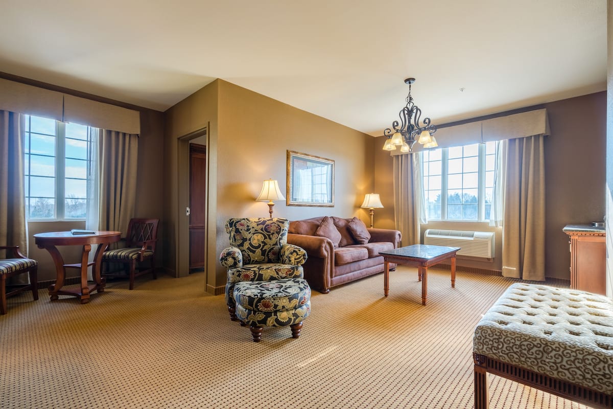 A spacious living room with a large window, a brown leather sofa, a patterned armchair, a wooden coffee table, and a chandelier.