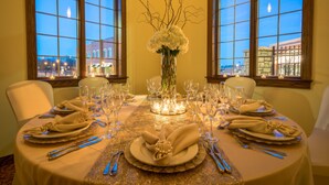 Une table à manger élégante, dressée pour six personnes, agrémentée d’un vase de fleurs blanches, de bougies et offrant une vue sur les bâtiments à travers de grandes fenêtres.