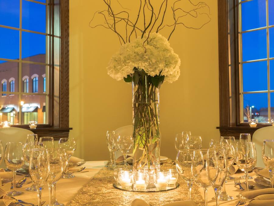 A table set for a formal dinner with a vase of flowers, candles, and a view of the cityscape through large windows.