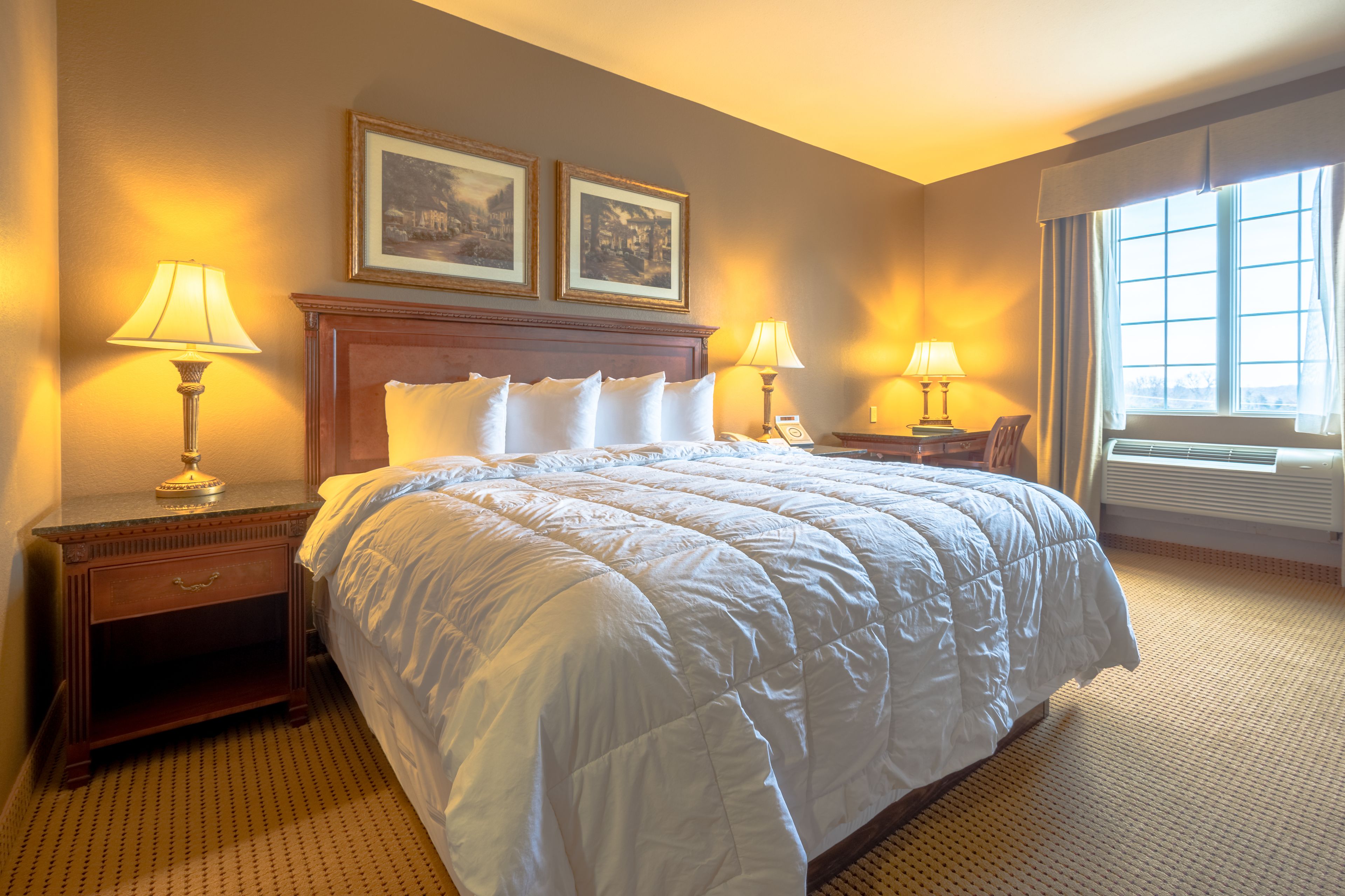 A hotel room with a large bed, two bedside tables with lamps, a wooden headboard, and two framed pictures on the wall.