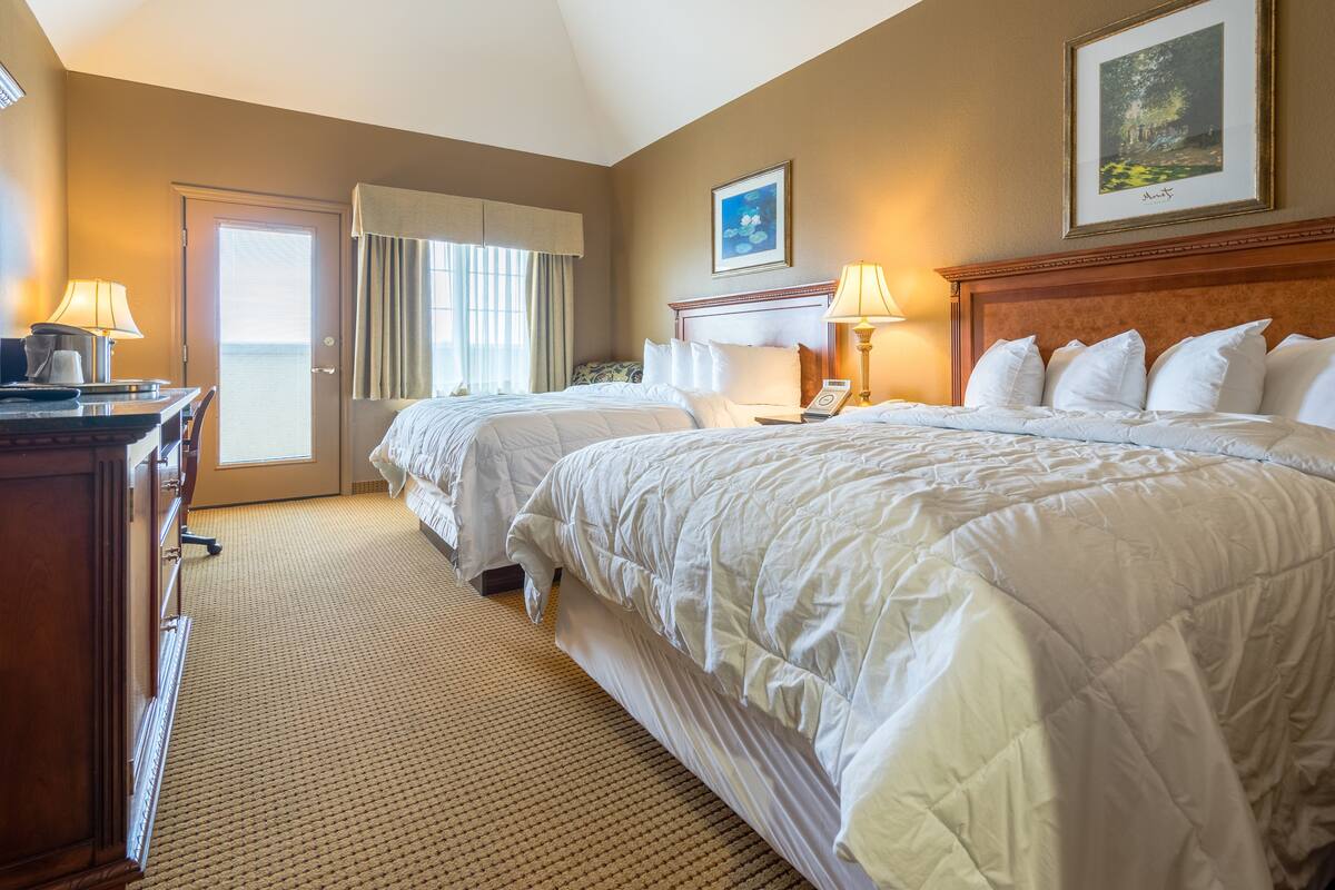 A hotel room with two beds, a desk, a chair, a lamp, a painting on the wall, and a door with curtains.