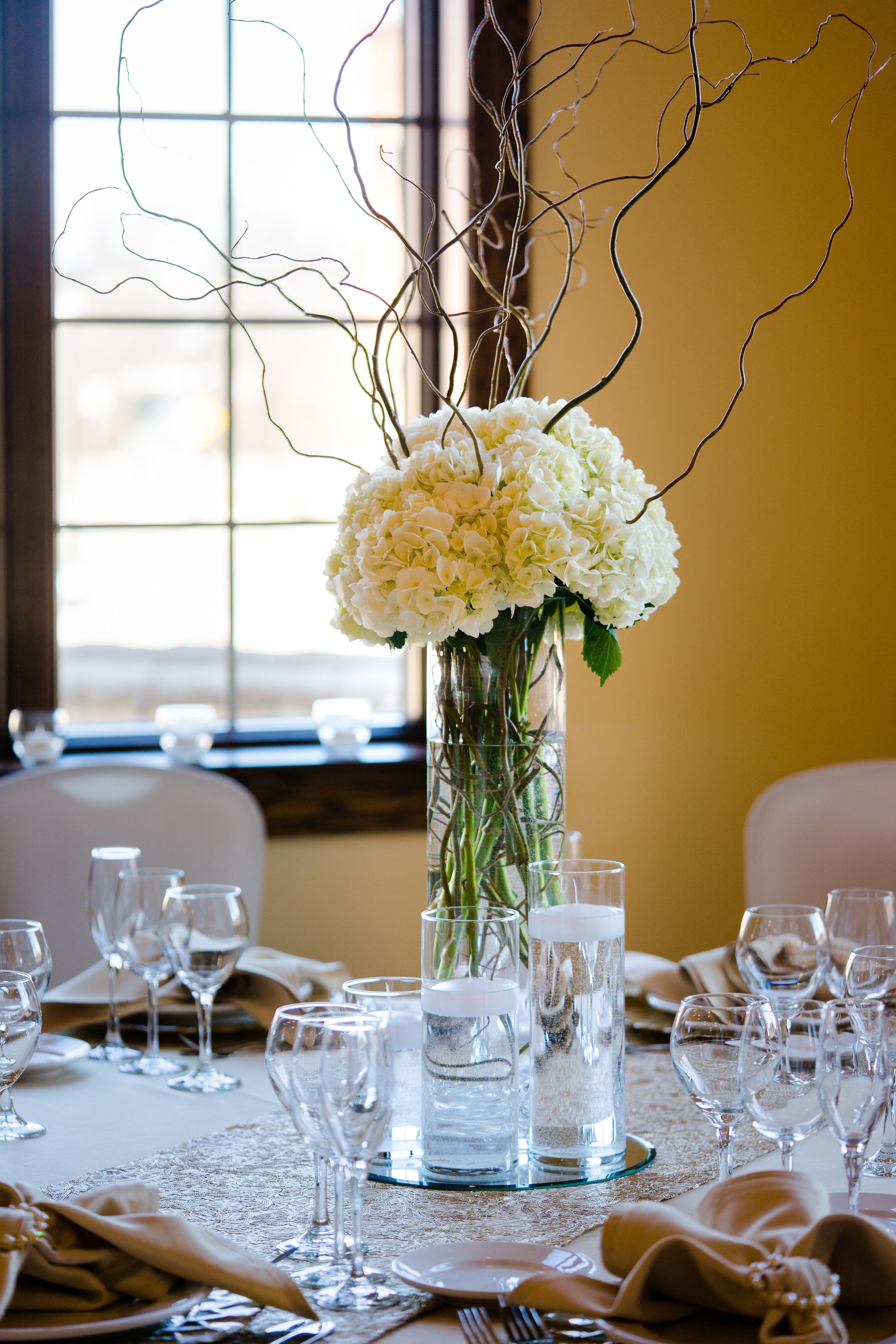 A table set for a formal event with a tall vase of white flowers, clear glasses, and a lace tablecloth.