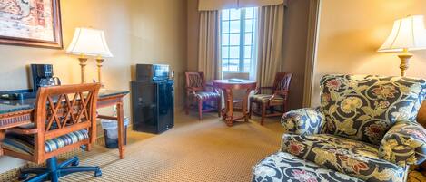 A hotel room with a desk, two chairs, a sofa, a small table, a TV, and a window with curtains.