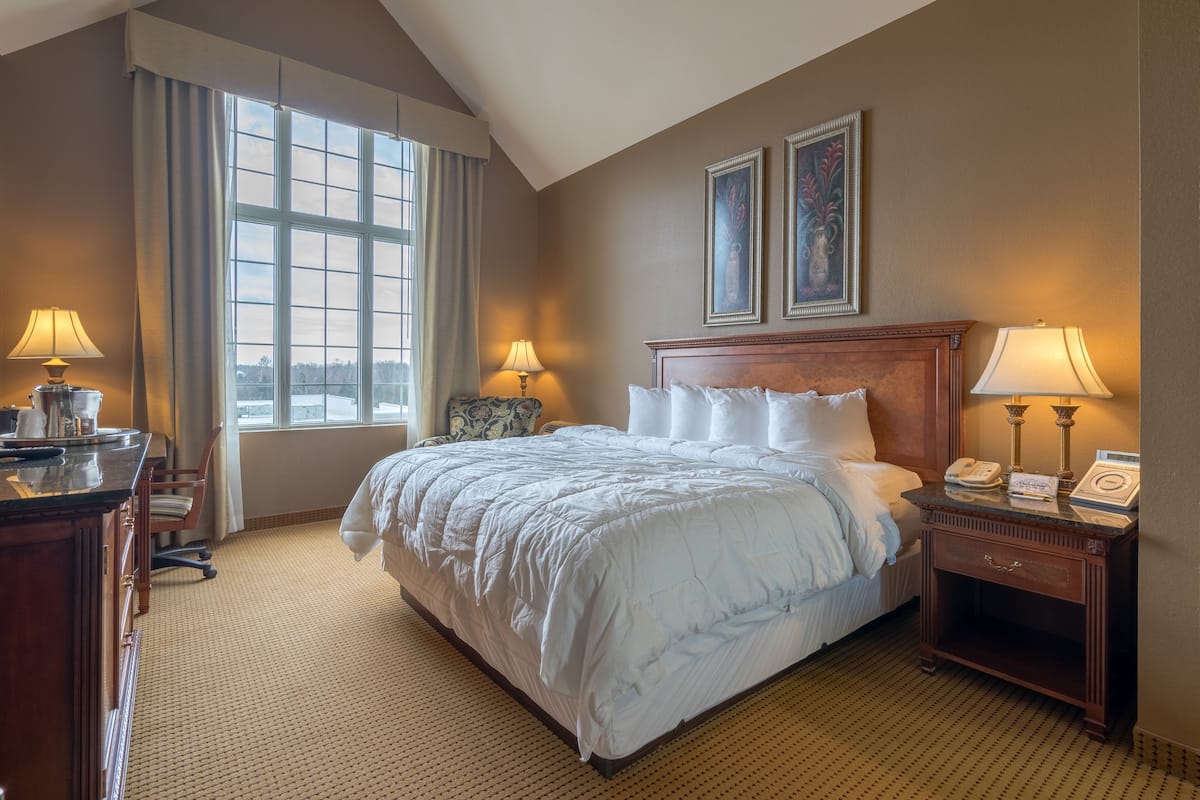 A hotel room with a large bed, two bedside lamps, a wooden nightstand, a desk with a lamp, and a window with curtains.