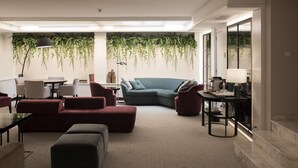 En moderne stue med en hjørnesofa, et sofabord og et spiseområde med stole. Der er hængende planter på væggen, en lampe og udsigt gennem store vinduer.