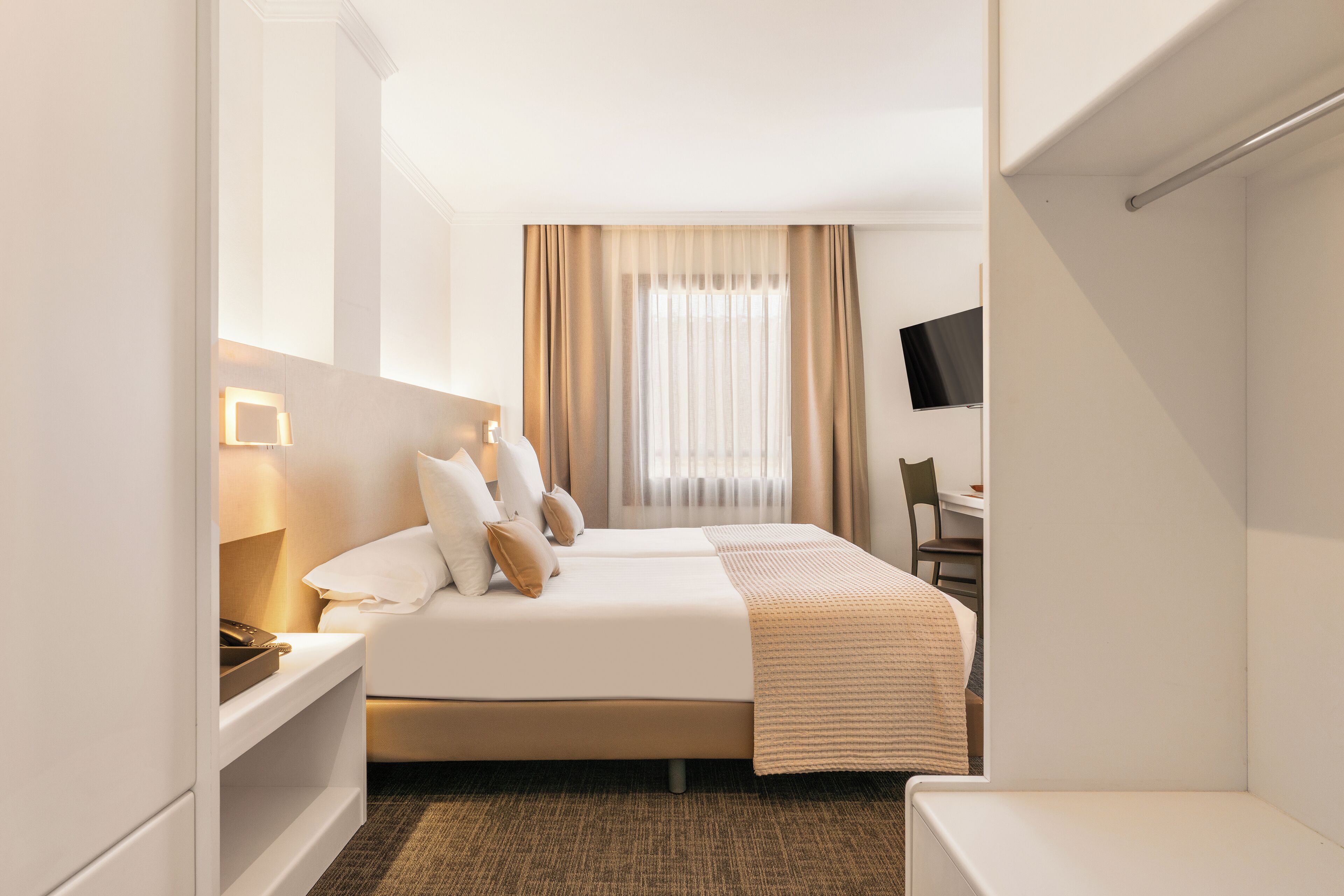 A hotel room with a bed, bedside tables, a desk, a chair, a television, and a window with curtains.