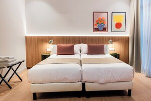 A double bed with two pillows, a nightstand with a lamp, a folded towel on a stand, and artwork on the wall.