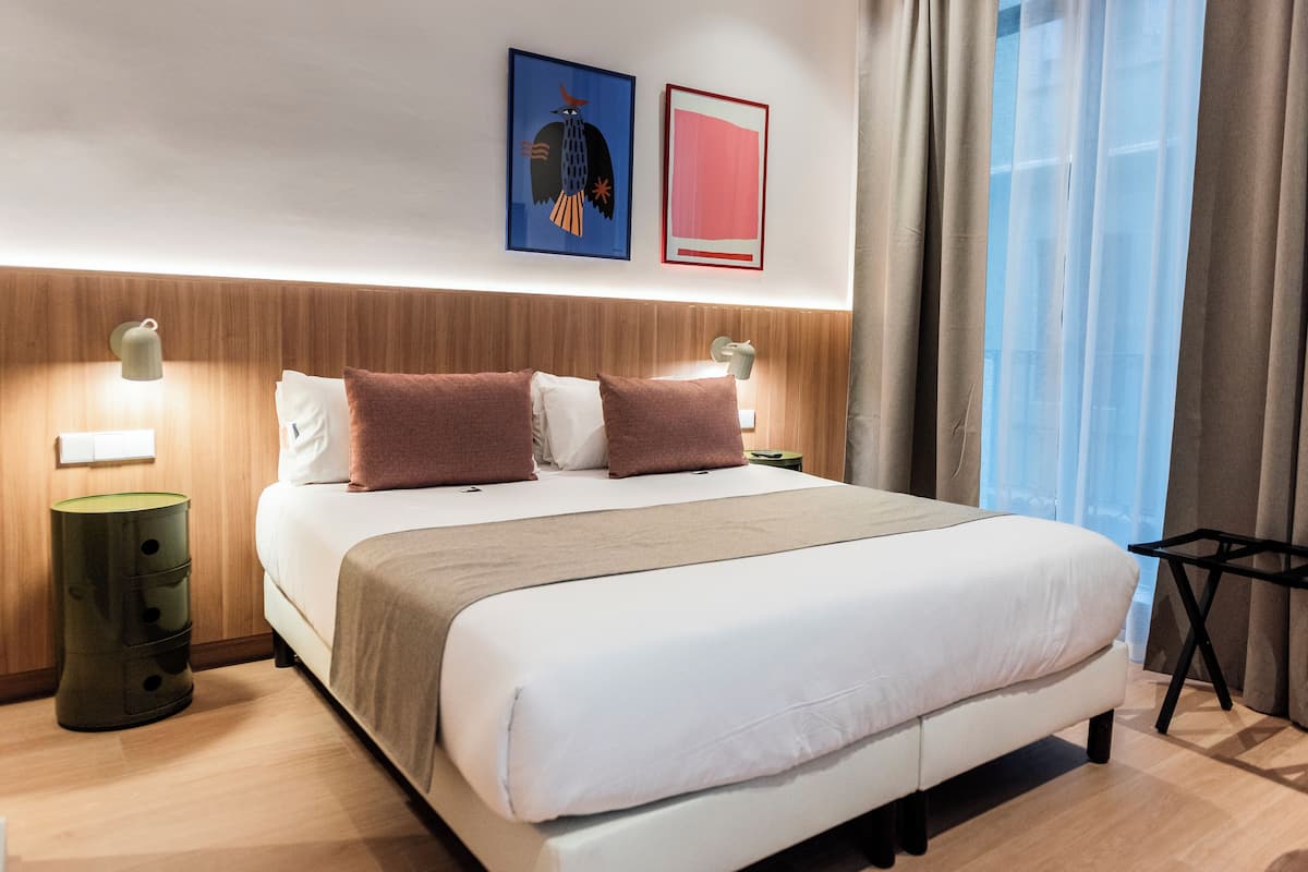 A neatly made bed with a white and beige duvet, two pillows, and a wooden headboard. There are two bedside tables with lamps, a green cylindrical nightstand, and a black side table.