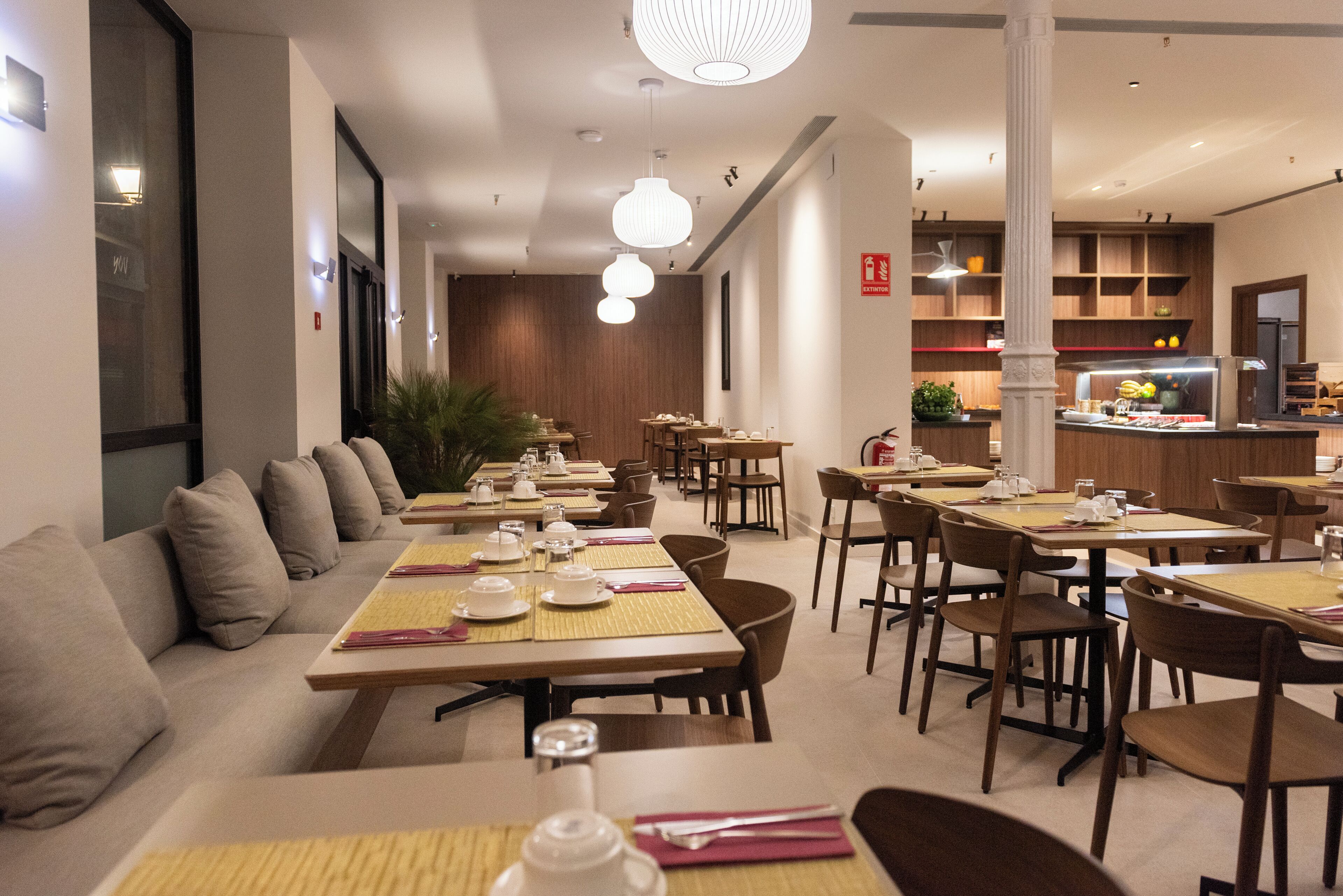 A modern restaurant with multiple dining tables and chairs, a long bench seating area, and a kitchen in the background.