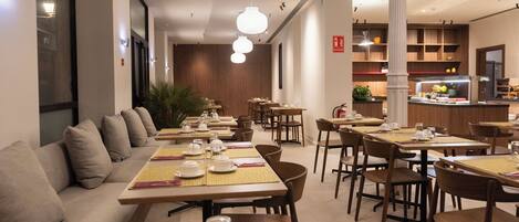 A modern restaurant with multiple dining tables and chairs, a long bench seating area, and a kitchen in the background.