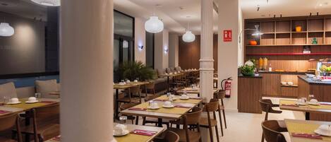 A restaurant with multiple dining tables, chairs, pendant lights, and a kitchen area.