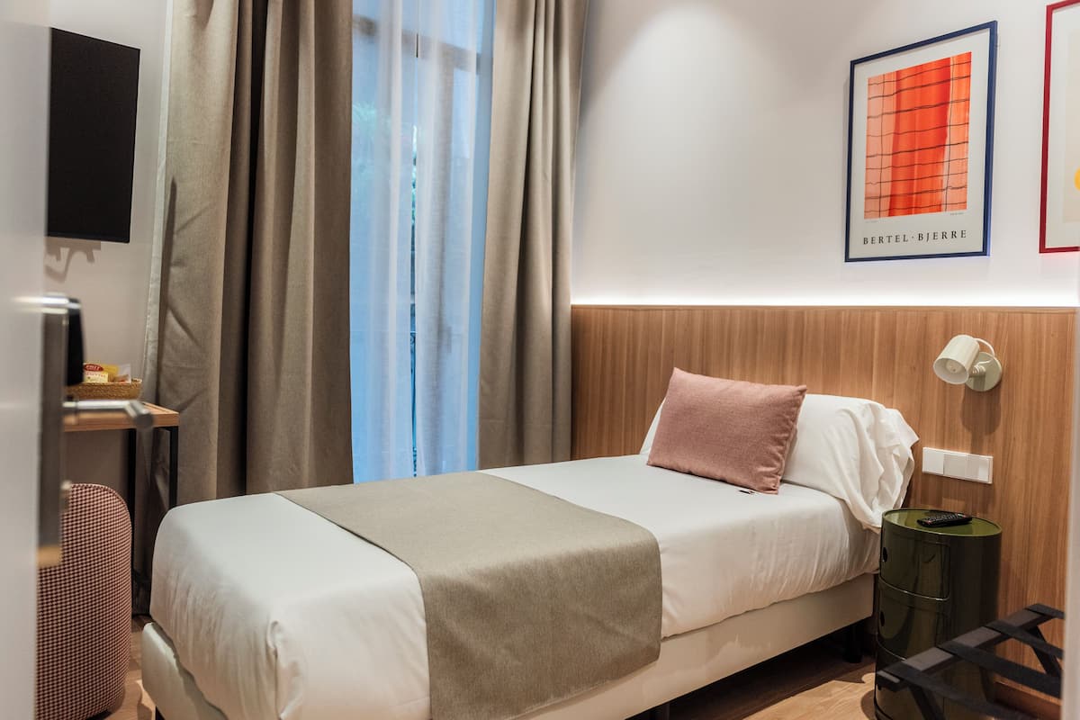 A hotel room with a bed, a bedside table, a lamp, a TV mounted on the wall, and a window with curtains.