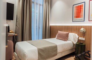 Habitación de hotel con cama, mesita de noche, lámpara, televisor montado en la pared y ventana con cortinas.