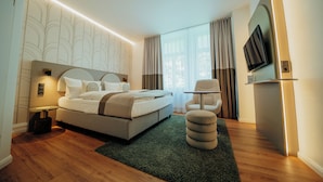 A modern hotel room with a bed, bedside table, lamp, chair, ottoman, and a flat-screen TV mounted on the wall. - guestified. BARBAROSSA Hotel (Ratingen)