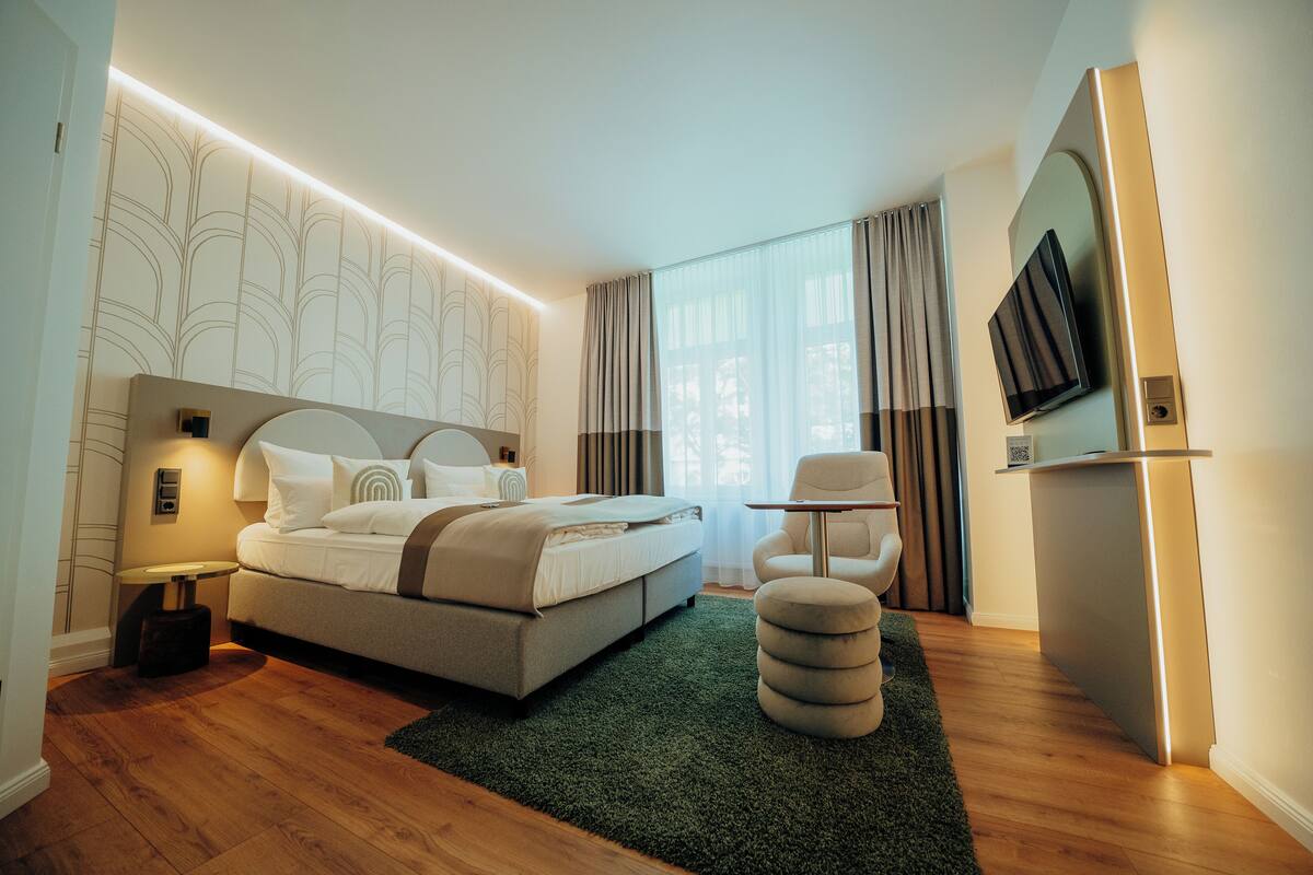 Een moderne hotelkamer met een bed, nachtkastje, lamp, stoel, pouf en een wandgemonteerde flatscreen-televisie.