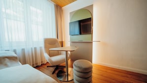 Une chambre d’hôtel avec un lit, un pouf rond, une petite table, une chaise et un téléviseur fixé au mur.