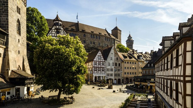 A cobblestone square with traditional half-timbered buildings, a prominent tower, and a tree providing shade. - Hotel Metropol Nürnberg (Nuremberg)
