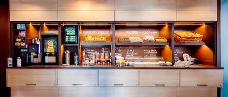 Una barra de desayuno con diversos alimentos y bebidas, que incluye una máquina de café y una presentación de frutas.